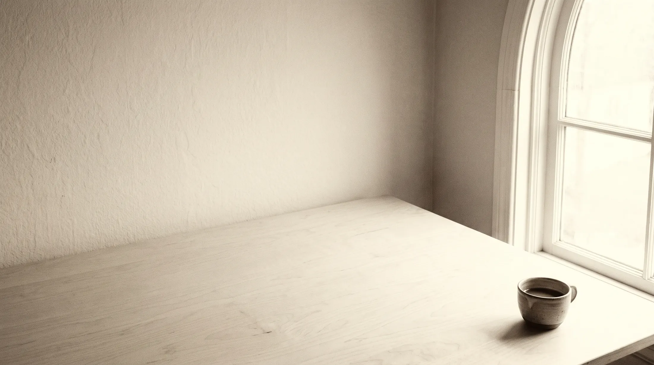 Ein leerer heller Schreibtisch am Fenster im Morgenlicht, darauf eine einzelne Tasse